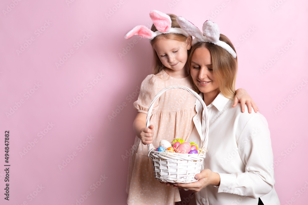Happy Easter. Portrait of beautiful blonde mother and daughter with rabbit ears and a basket of colored eggs in their hands. Pink background. Space for text.