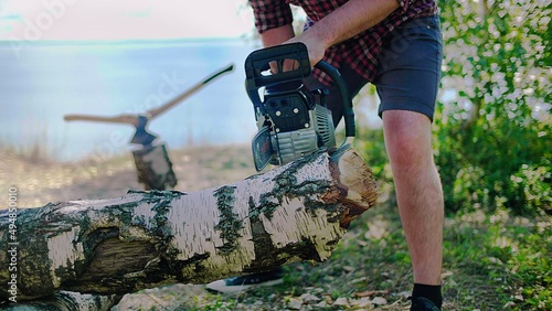 A woodcutter, logger saws a wood with a chainsaw on the beach of a lake, river