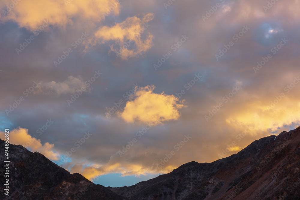 Scenic landscape with dark mountain range silhouette under evening blue sky with vivid orange sunset clouds. Sunlit orange cirrus clouds in sunset sky above mountains silhouettes at changeable weather