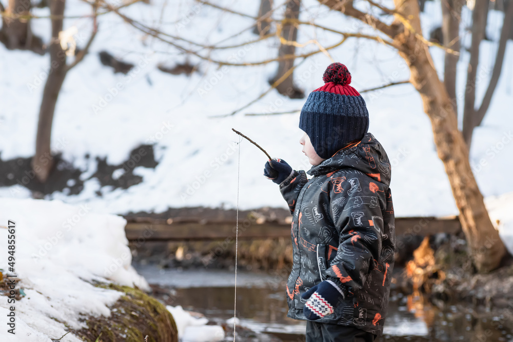 Fototapeta premium A little boy in the park in early spring. A child is playing with a homemade fishing rod made of a stick. Wooden bridge over the stream.