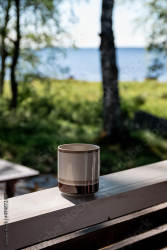 Grey and brown coffee mug on a wooden railing with blurred background of nature and lake in summer in Finland
