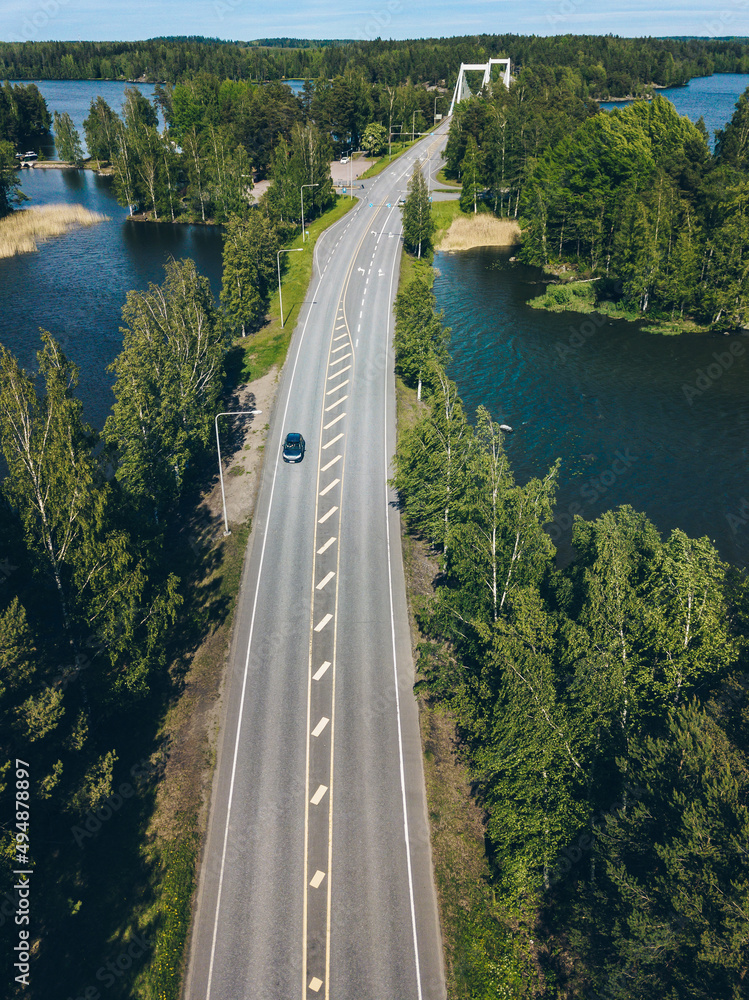 Obraz premium Aerial view of road through blue lake and green woods in Finland.