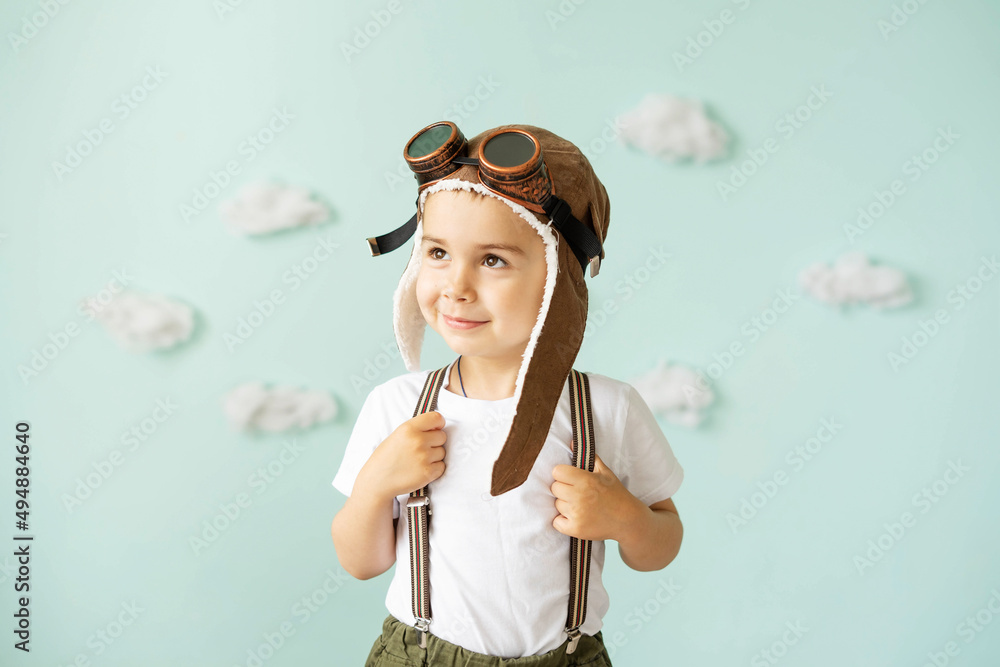 A boy in an aviator helmet and suspenders stands on a blue background with clouds and smiles. A boy in an aviator helmet and suspenders stands on a blue background with clouds and smiles.
