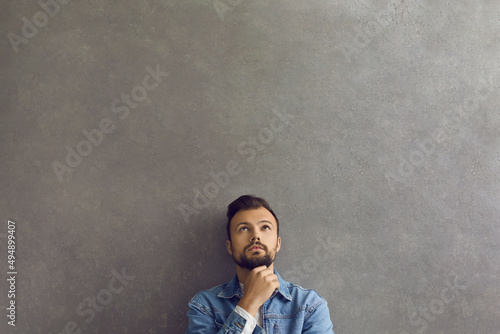 Serious handsome man in modern casual denim jacket leaning on wall, thinking and looking up at copy space above head. Young guy dreaming about something standing on grey text space background photo