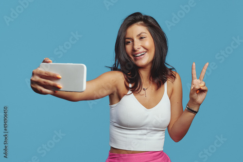 Stylish cheerful young woman showing peace gesture while having video-call, holding smart phone, wearing white top, isolated on blue background photo