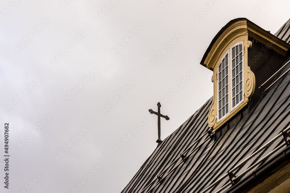 Metal cross at the rooftop against grey cloudy sky at the roof of the building. Detail of the church roof with cross and decorated roof window. Cross protecting the sky on roof.
