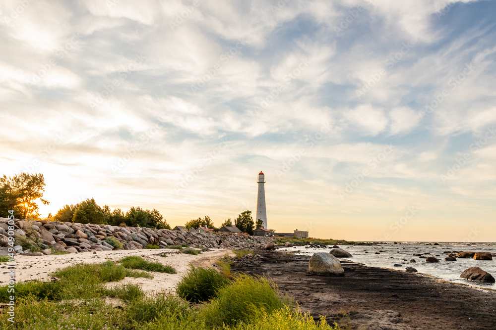 Obraz premium Coastal sunset near Tahkuna lighthouse at Hiiumaa, Estonia, Europe. White lighthouse with red top on the rocky beach at dusk. Sun is setting on the shore near lighthouse.