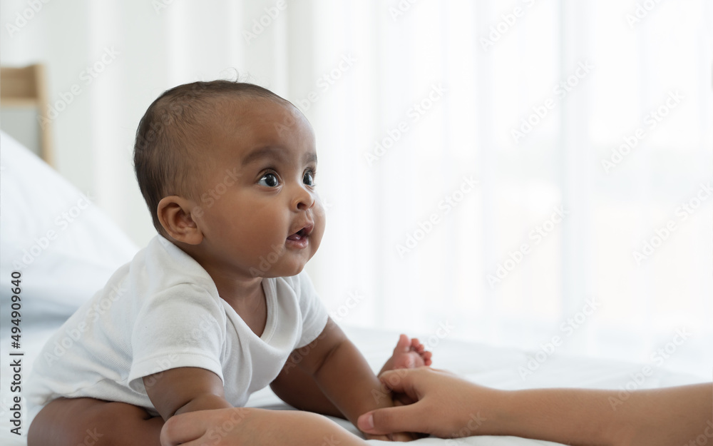Portrait of little adorable African newborn baby sitting on bed and looking with bright eyes and smiling and holding mother hands at home. Motherhood, child care concept Portrait of little adorable African newborn baby sitting on bed and looking with bright eyes and smiling and holding mother hands at home. Motherhood, child care concept