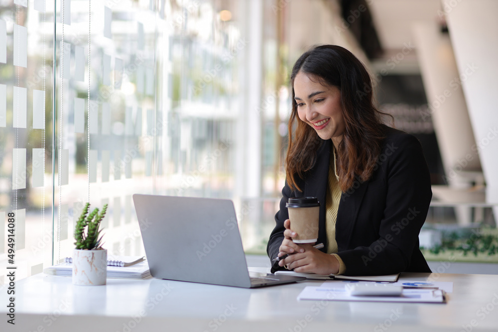Attractive Asian secretary or businesswoman holding hot coffee mug sitting on laptop in office.
