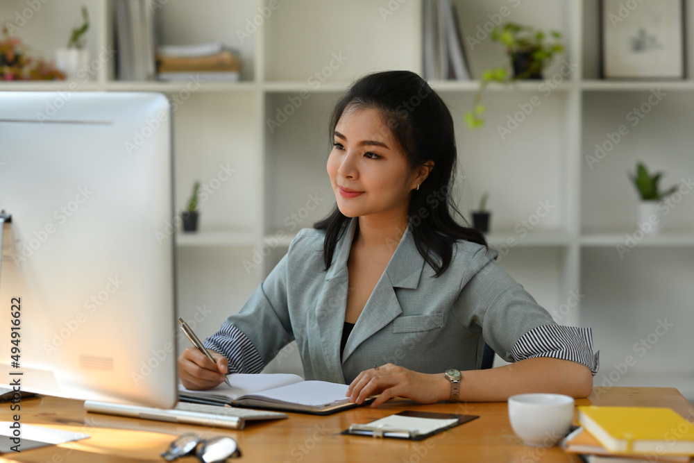 A portrait of a pretty Asian woman sitting in the office looking at a computer and working on a note, for business and technology concept. A portrait of a pretty Asian woman sitting in the office looking at a computer and working on a note, for business and technology concept.