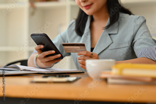 Cropped view of a pretty smiling Asian woman sitting in the office holding a credit card and a smart phone, for finance and technology concept.