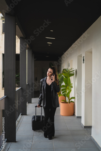 Chica joven con pelo largo vestida de negro con maleta de viaje con ruedas por pasillo de hotel estilo soho © MiguelAngelJunquera