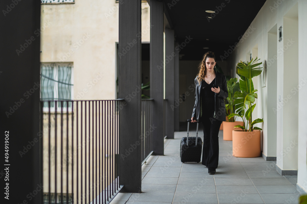 Chica joven con pelo largo vestida de negro con maleta de viaje con ruedas por pasillo de hotel estilo soho Chica joven con pelo largo vestida de negro con maleta de viaje con ruedas por pasillo de hotel estilo soho