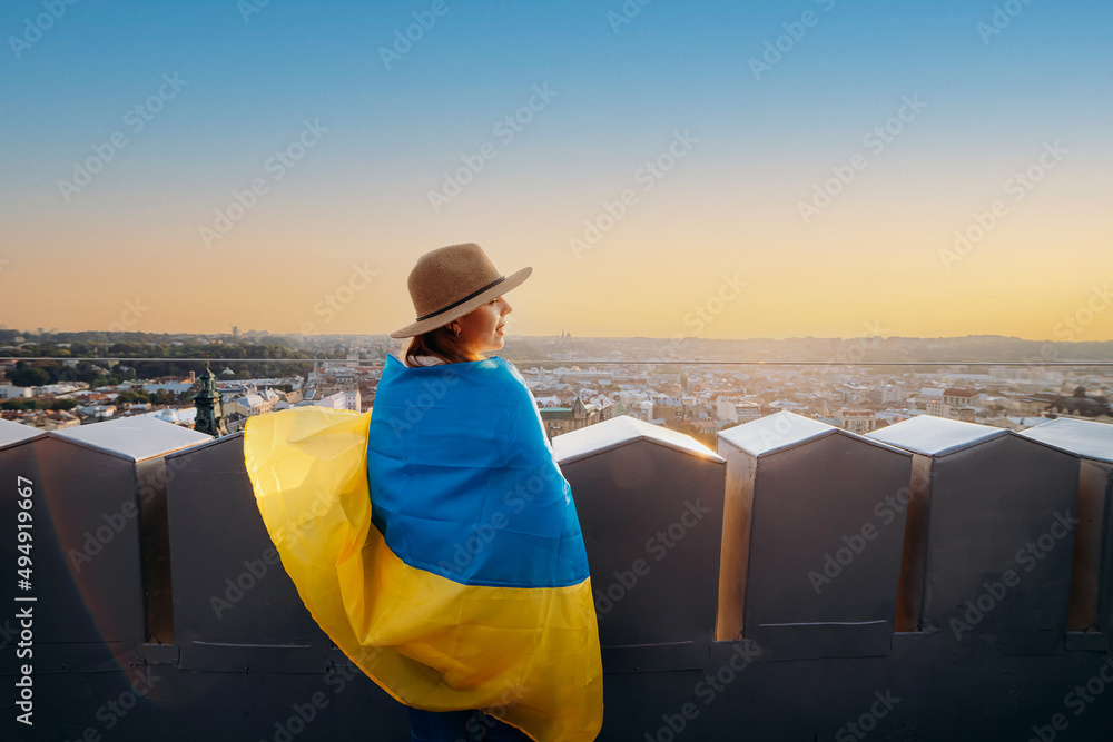 Fototapeta premium A woman stands with the national Ukrainian flag and waving it praying for peace at sunset in Lviv..A symbol of the Ukrainian people, independence and strength. Pray for Ukraine.