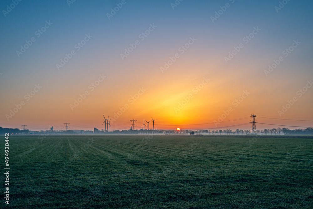 Panoramic image of the sunrise over a Dutch rural landscape in the province of North Brabant. In the background the silhouettes of wind turbines and high voltage pylons and lines are visible.