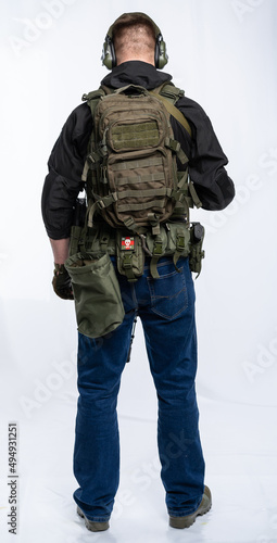 airsoft player in full growth. a man in an outfit, in headphones, a bulletproof vest, with a rifle and a pistol on a white background. standing with his back to us