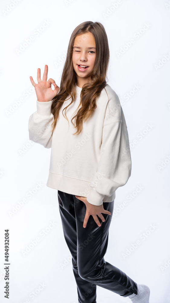 portrait of a girl in the studio. beautiful little girl in a white sweater and trousers smiles, points ok and winks. isolated white background portrait of a girl in the studio. beautiful little girl in a white sweater and trousers smiles, points ok and winks. isolated white background