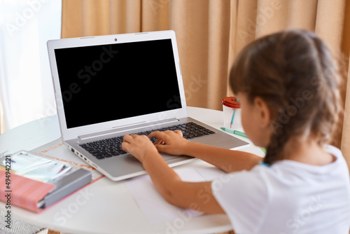 Portrait of little girl with braids having online lesson during distance education, sitting on table and typing on keyboard, display with space for promotional text, distance education.