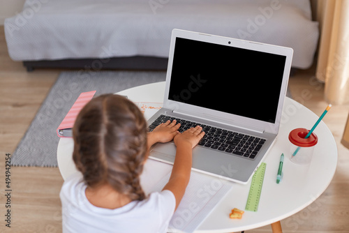 Back view of dark haired little girl having online lesson during distance education, sitting on table and typing, distance education, learning process.