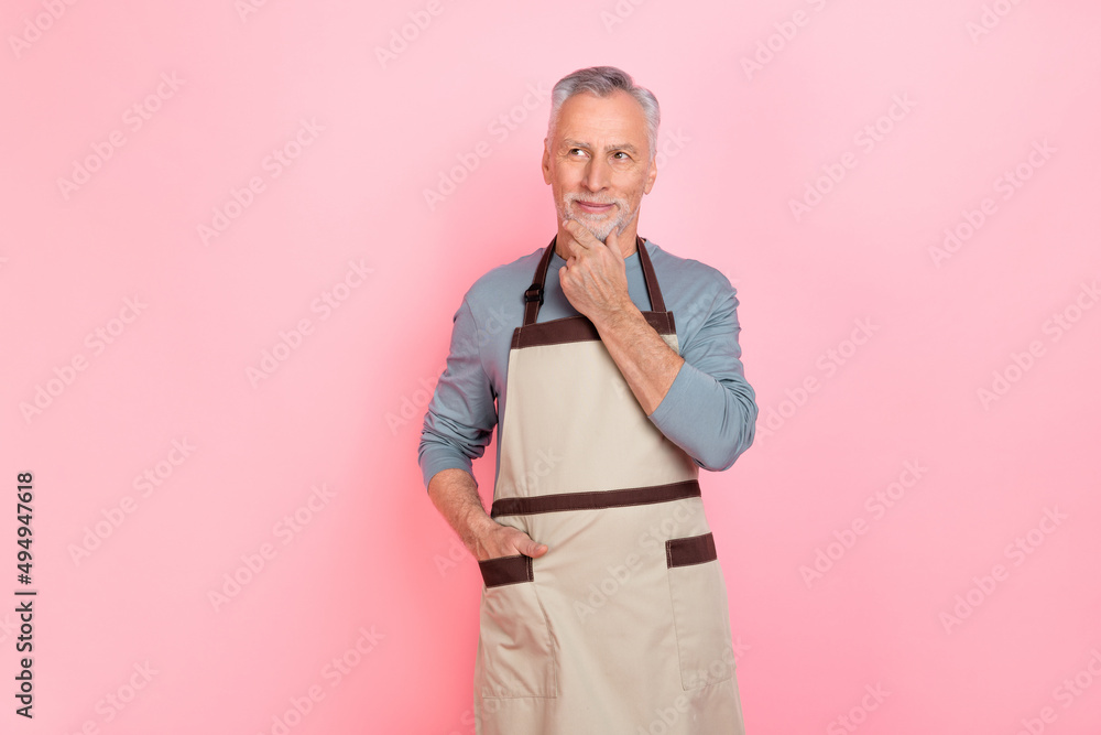 Photo of charming positive retired man wear grey shirt apron hand arm chin looking empty space isolated pink color background Photo of charming positive retired man wear grey shirt apron hand arm chin looking empty space isolated pink color background