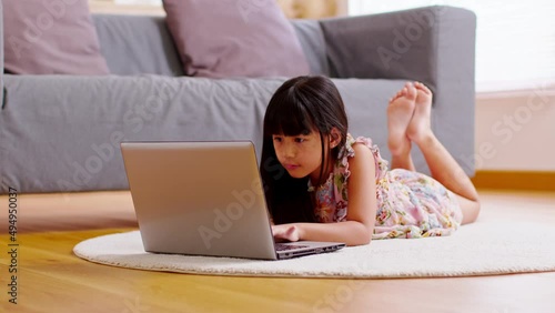 Cute little girl uses laptop while prone at the sofa in the living room. Child surfing the internet on computer, browses through internet and watches cartoons online. photo