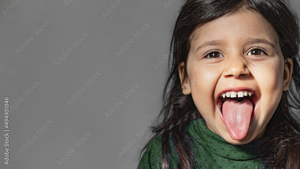 A pretty girl with dark hair and eyes sticks out her tongue on a gray background. Close up portrait of emotional preschooler in green longsleeve A pretty girl with dark hair and eyes sticks out her tongue on a gray background. Close up portrait of emotional preschooler in green longsleeve