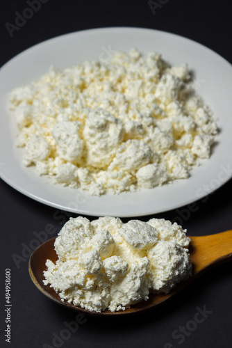 A plate of cottage cheese on a black background. Healthy and tasty dairy product. Wooden spoon full of cottage cheese