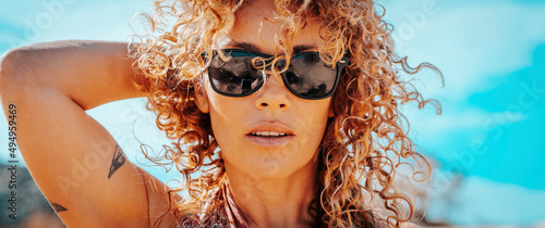 Summer portrait of beautiful young adult woman with sunglasses and healthy blonde long curly hair looking at camera. Attractive brunette with blue sky in background. One cute female people lifestyle