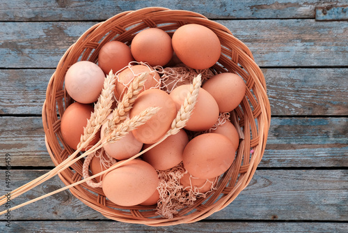 Brown beautiful eggs in a light wooden basket with wheat ears on top on a blue table background. The concept of farming, dietary product, cooking.
