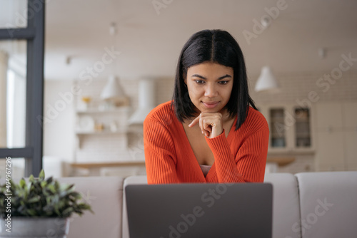 Beautiful African American businesswoman using laptop computer watching training courses working online from home. Pensive female shopping online, ordering food. Student studying, learning language photo