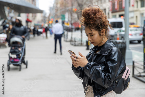 New York City lifestyle featuring young woman with curly hair wearing black leather jacket outside looking at mobile phone