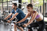 Young sporty couple doing cardio work out on stationery exercise bikes in indoor fitness gym. Man and woman exercising and cycling bikes together in gym. Healthy lifestyle concept.