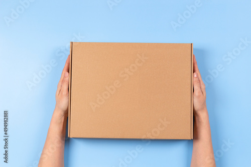 Top view to female hands holding brown cardboard box on light blue background. Mockup parcel box. Packaging, shopping, delivery concept © vejaa