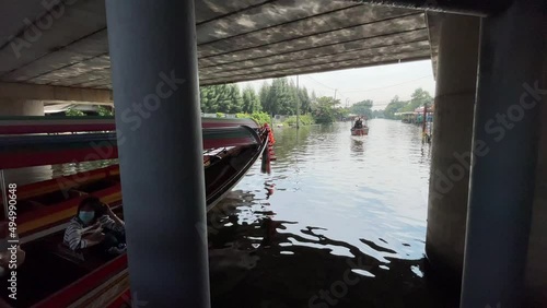 BANGKOK, THAILAND - Circa November, 2021: Long tail boat parks at Taling Chan Floating Market Pier. Passengers inside the tour boat floating on a weekend market under the bridge on a sunny day photo