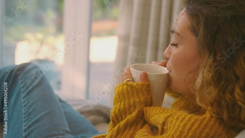 Young woman in cosy warm jumper sitting in chair at home looking out of window with hot drink - shot in slow motion
