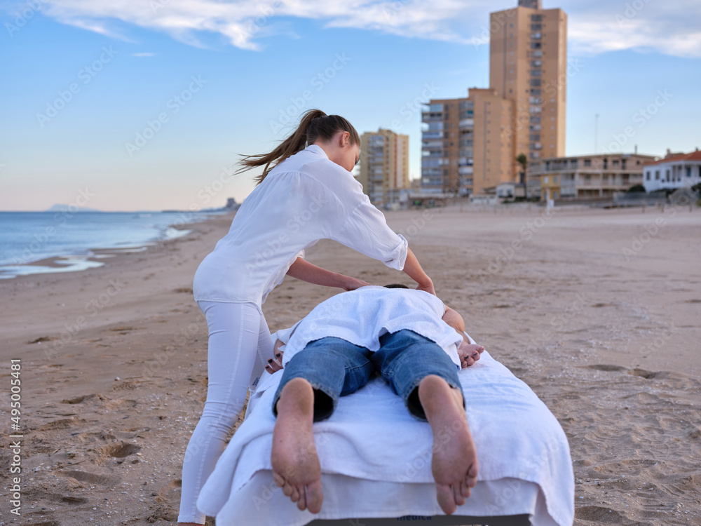 Fototapeta premium A young chiromassage therapist giving a massage to young adult man to treat his back injury on a beach in Valencia with buildings on background.