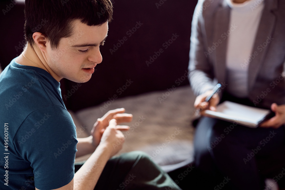 Shy down syndrome man during conversation with psychologist at home. - obrazy, fototapety, plakaty 