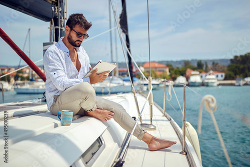 A young man watching a tablet content while sitting on a yacht and riding through the dock on the seaside. Summer, sea, vacation