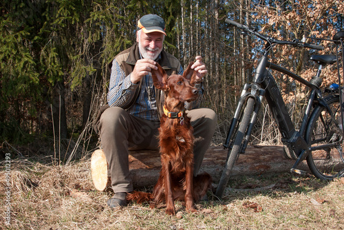 Dogs make people happy. Cyclist sits at the edge of the forest and has fun with his dog. He holds up his Irish Setter's floppy ears and laughs heartily.
