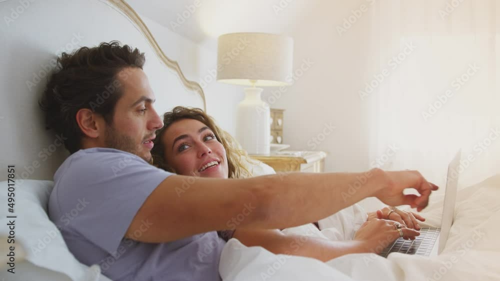 Loving couple wearing pyjamas lying in bed at home together looking at laptop - shot in slow motion Loving couple wearing pyjamas lying in bed at home together looking at laptop - shot in slow motion