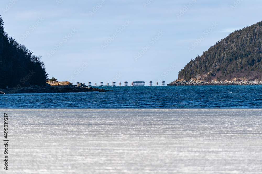 Obraz premium A wide view of frozen ice or slob in a cove. This ice has patches of snow with a glass surface. There are mountains or hills in the distance that are covered in green evergreen trees with blue sky.