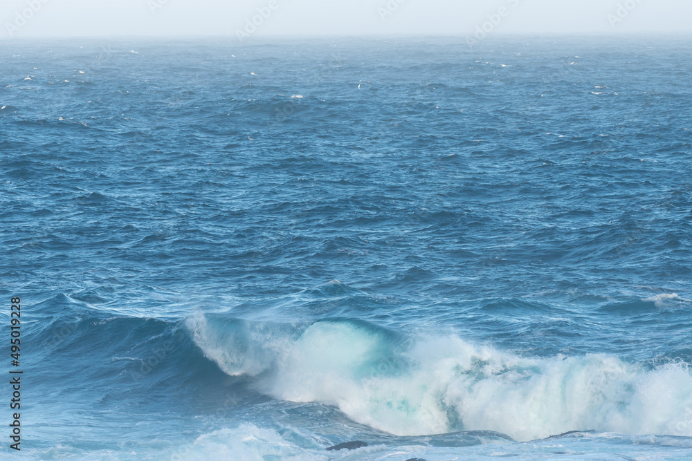 Obraz premium A large blue colored rip wave rolling along the beach in the cold North Atlantic Ocean. There's spray coming off the foam along the rip of the wave. The background is a dark majestic blue sea.