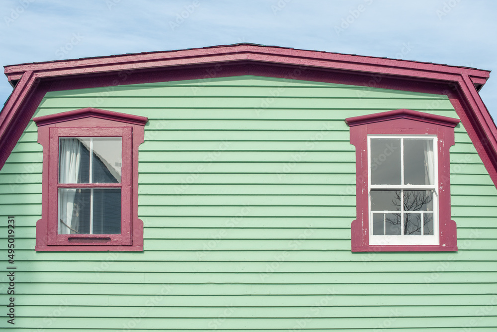 A vibrant lime green exterior wall of narrow horizontal cape cod clapboard on a house. There are two single four pane glass casement windows with bright pink decorative trim with white lace curtains