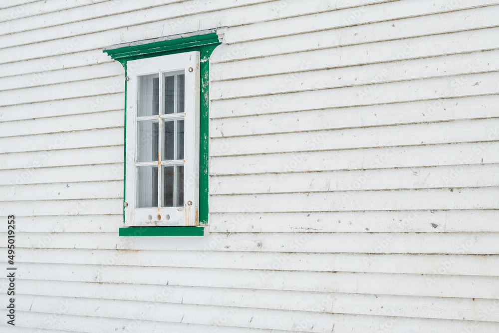 A small single casement window in a white narrow wood beveled clapboard siding exterior wall of a vintage house. The glass window has six panes with green decorative trim on the outside edge. A small single casement window in a white narrow wood beveled clapboard siding exterior wall of a vintage house. The glass window has six panes with green decorative trim on the outside edge.