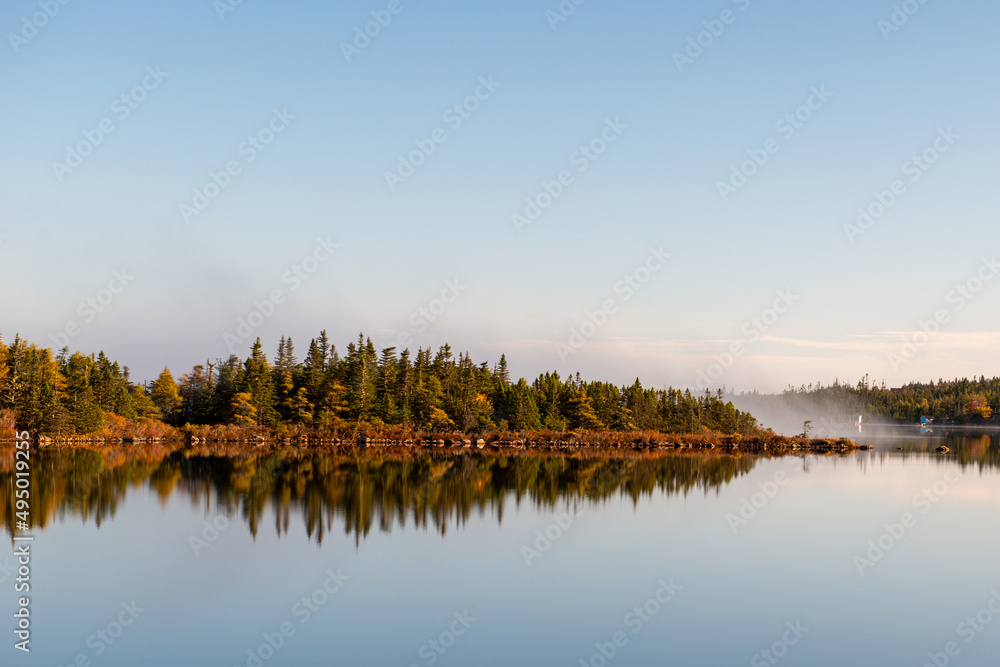 Fototapeta premium The edge of a pond with tall green and orange colored trees. The autumn forest and deep blue sky are reflected in the calm blue pond water. There's a light mist on the water along the riverbank.