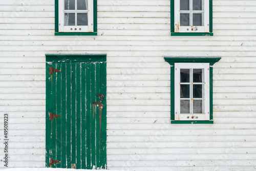 The exterior wall of a vintage white wooden building with bright green trim. There's a small wooden door and three glass windows in the country house. The wall has a textured horizontal clapboard.