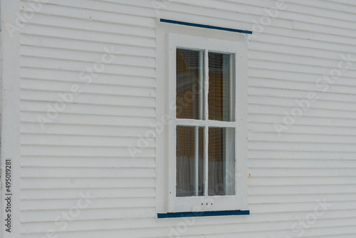 A small single casement window in a white narrow wood beveled clapboard siding exterior wall of a vintage house. The glass window has six panes with green decorative trim on the outside edge. 