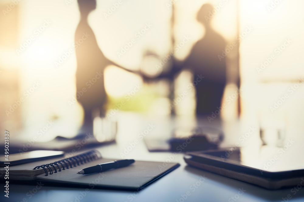 All for success in the name of business. Closeup shot of a notebook on a desk with businesspeople shaking hands in the background. All for success in the name of business. Closeup shot of a notebook on a desk with businesspeople shaking hands in the background.