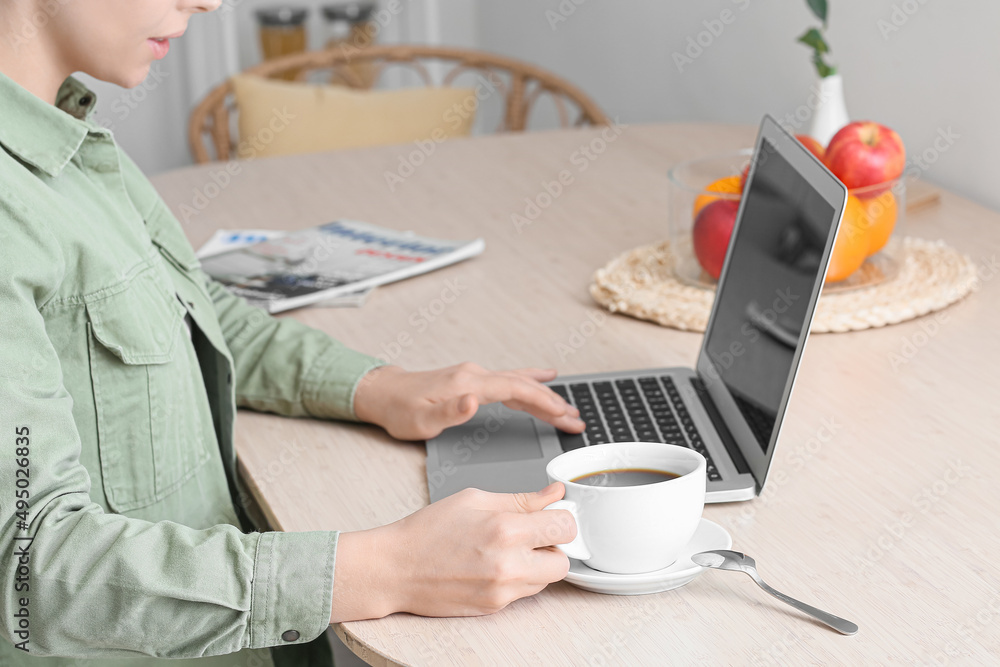 Young woman with cup of hot coffee using laptop at table in kitchen, closeup Young woman with cup of hot coffee using laptop at table in kitchen, closeup