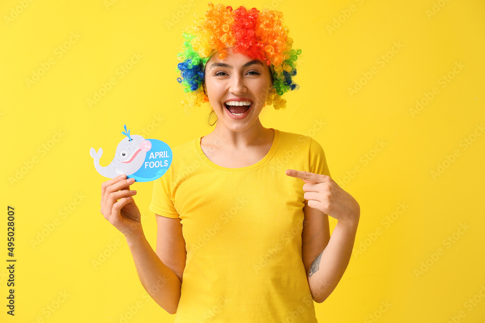 Funny young woman in colorful wig and with paper fish on yellow background. April fools' day celebration Funny young woman in colorful wig and with paper fish on yellow background. April fools' day celebration